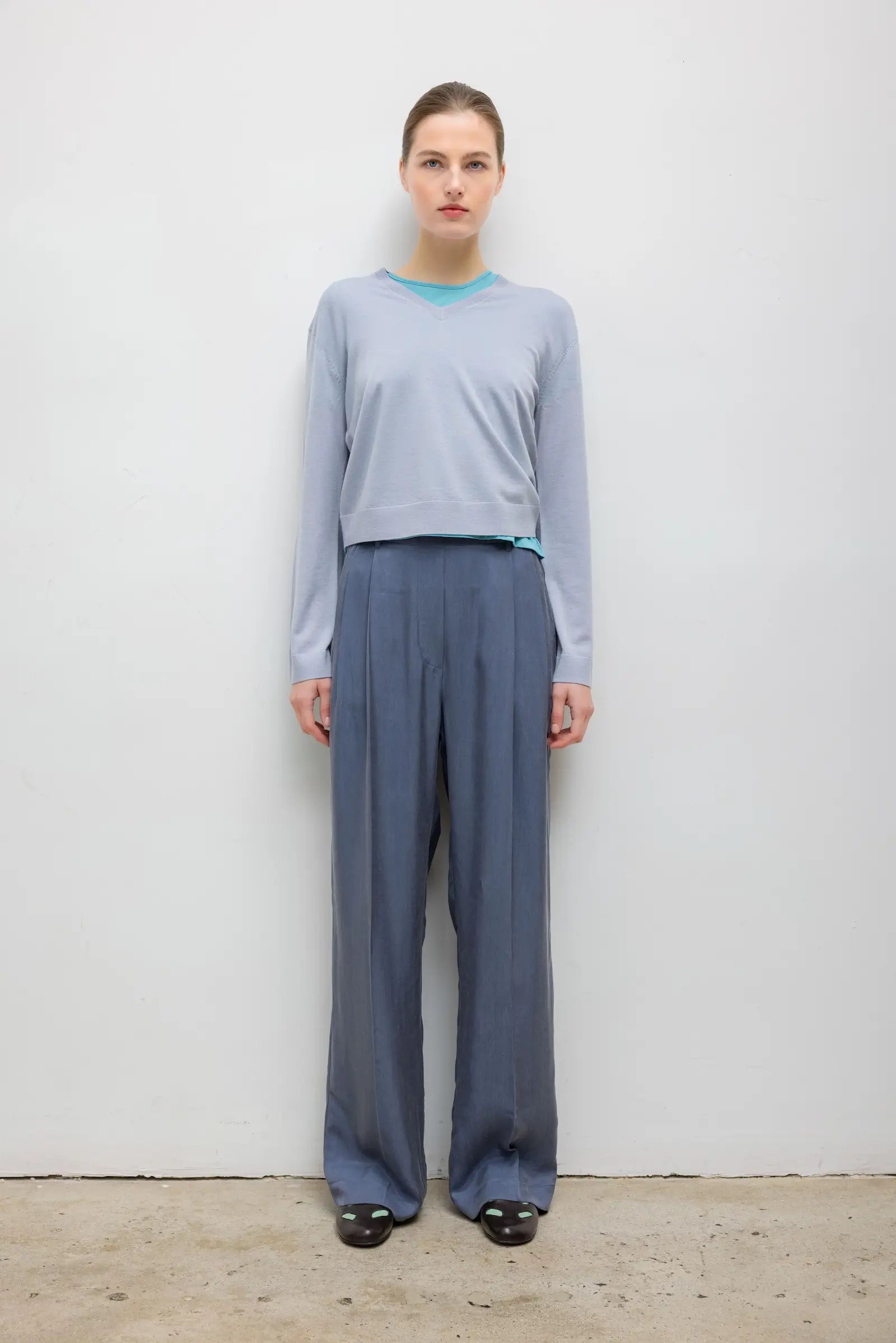 A model stands against a plain white wall wearing a light blue-gray long-sleeve crop top with a turquoise collar and matching wide-leg trousers in a muted blue-gray color.