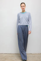 A woman modeling a light blue long-sleeve knit top and slate blue wide-leg trousers against a plain off-white wall.