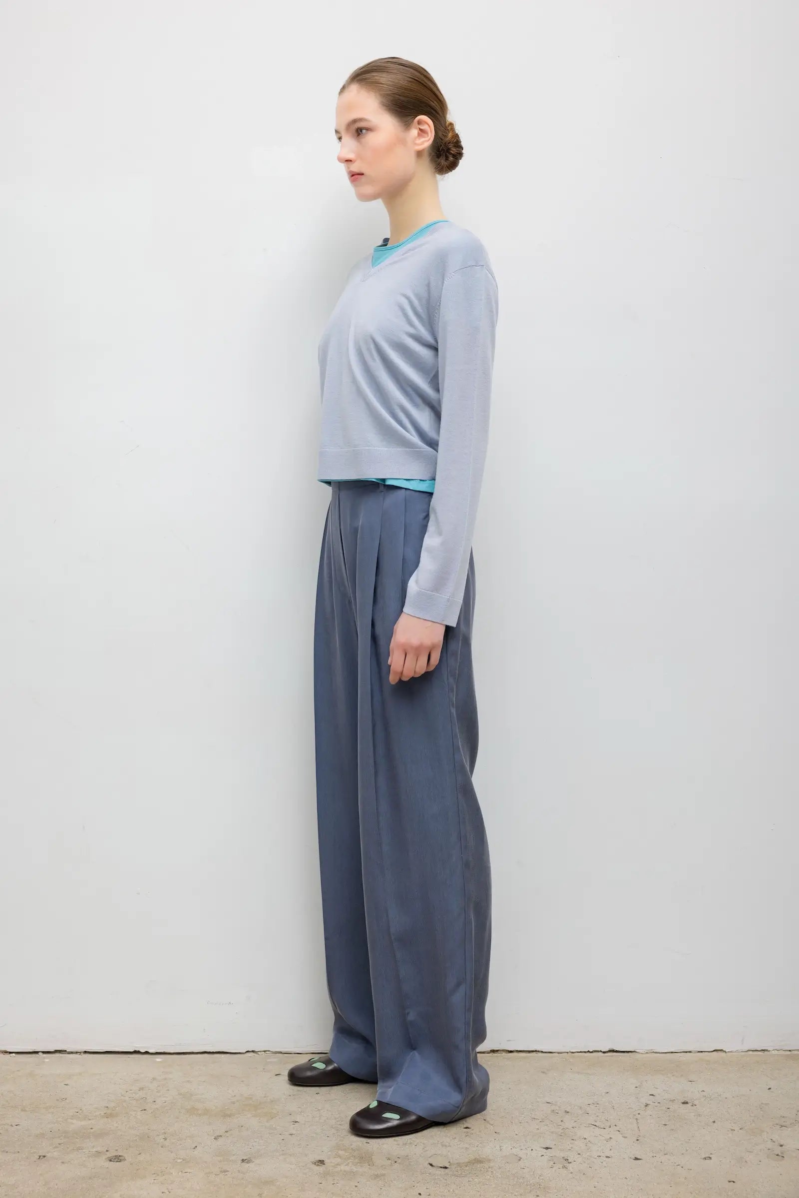 A woman stands in profile wearing a light blue long-sleeve cropped top, wide-leg slate blue pants, and black shoes against a plain off-white wall.