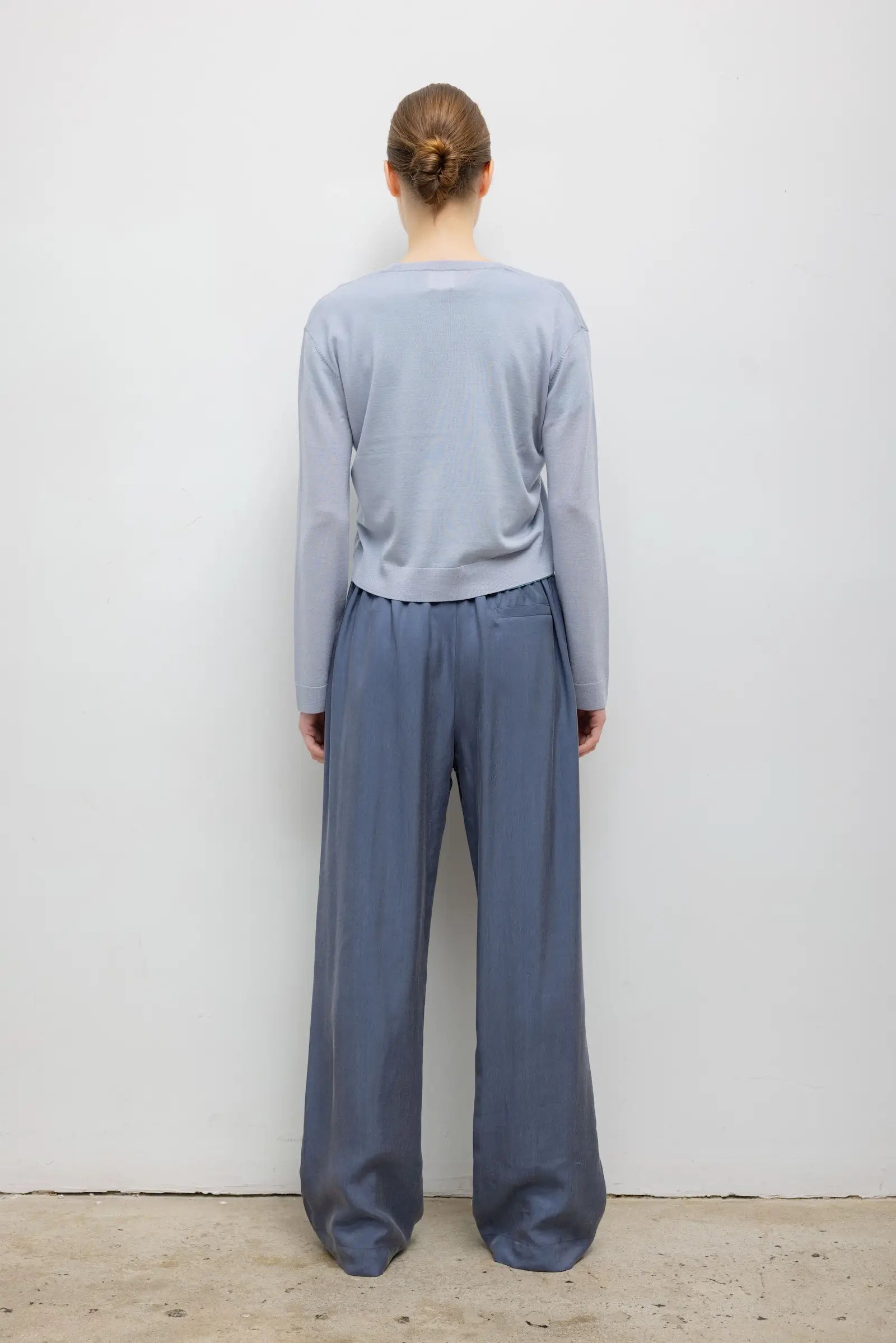 A woman modeling a light blue-gray long-sleeve cropped sweater with a bright turquoise inner collar and hem detail, paired with slate blue wide-leg trousers, against a plain light gray background from a back side view.