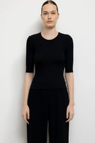 A woman modeling a black short-sleeve crewneck top and black wide-leg pants against a plain light gray studio background.
