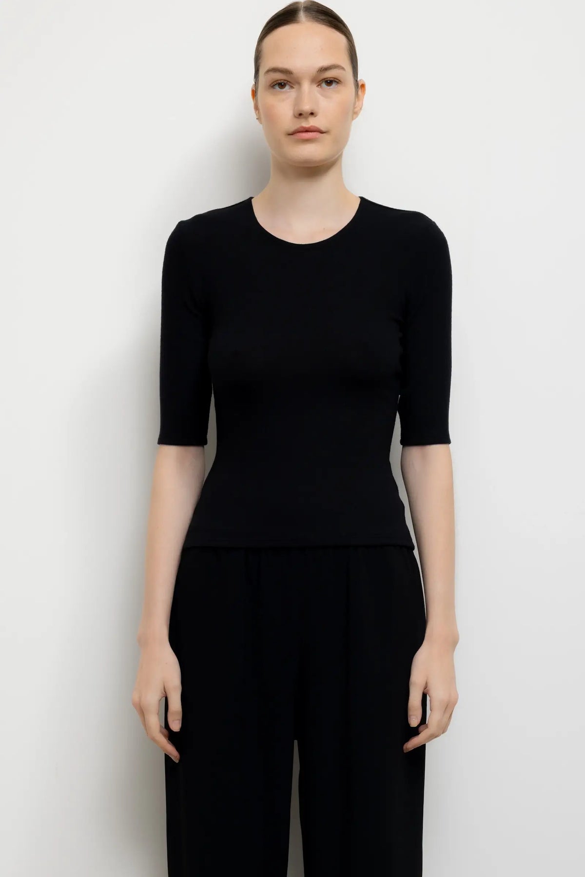 A woman modeling a black short-sleeve crewneck top and black wide-leg pants against a plain light gray studio background.
