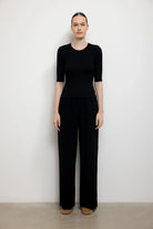 A model stands against a plain white wall wearing a black short-sleeve top and matching black wide-leg trousers, paired with tan shoes.