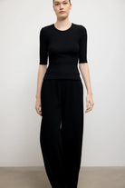 A model wearing a black three-quarter sleeve top and matching black wide-leg trousers, standing against a plain light gray studio wall.