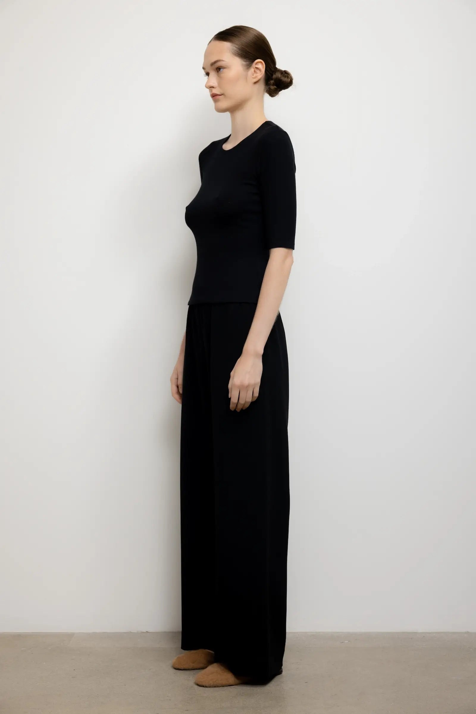 A woman stands in profile wearing a black short-sleeve knit top and matching black wide-leg pants against a plain light gray wall and beige floor.