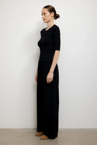A woman stands in profile wearing a black short-sleeve knit top and matching black wide-leg pants against a plain light gray wall and beige floor.