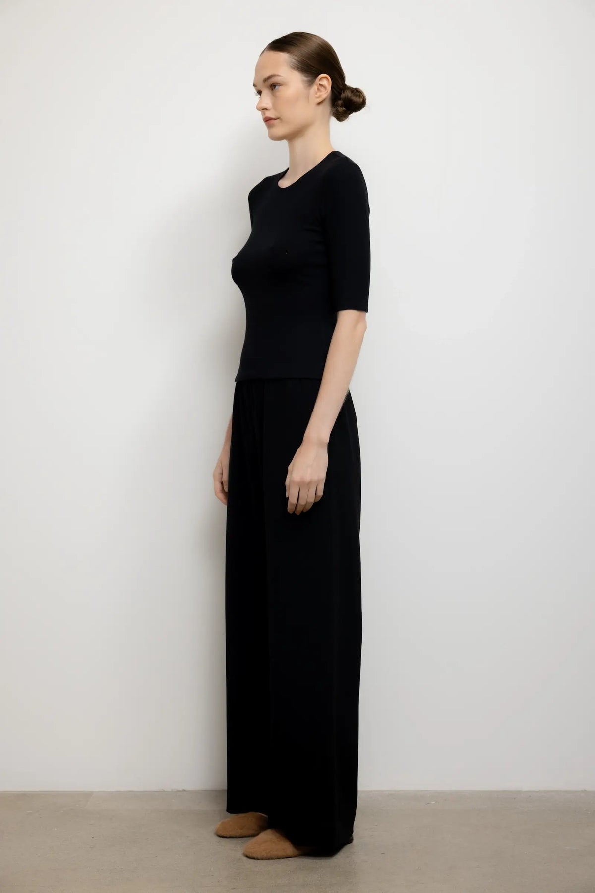A woman stands in profile wearing a black short-sleeve knit top and matching black wide-leg pants against a plain light gray wall and beige floor.