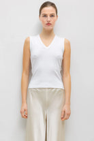 A woman modeling a white sleeveless V-neck top paired with light beige wide-leg pants, standing against a plain light gray studio background.