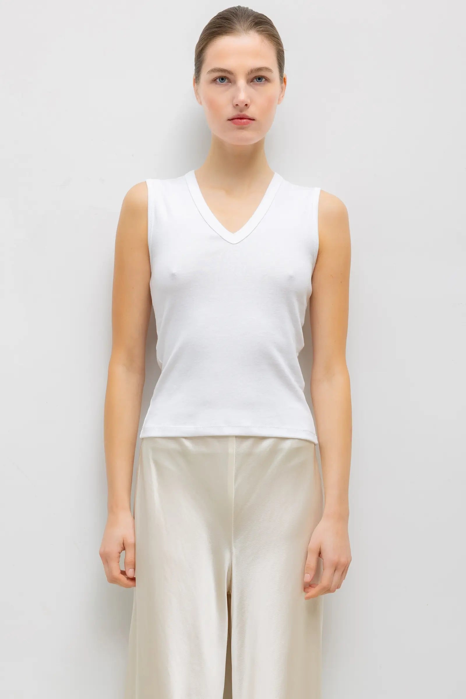 A woman modeling a white sleeveless V-neck top paired with light beige wide-leg pants, standing against a plain light gray studio background.