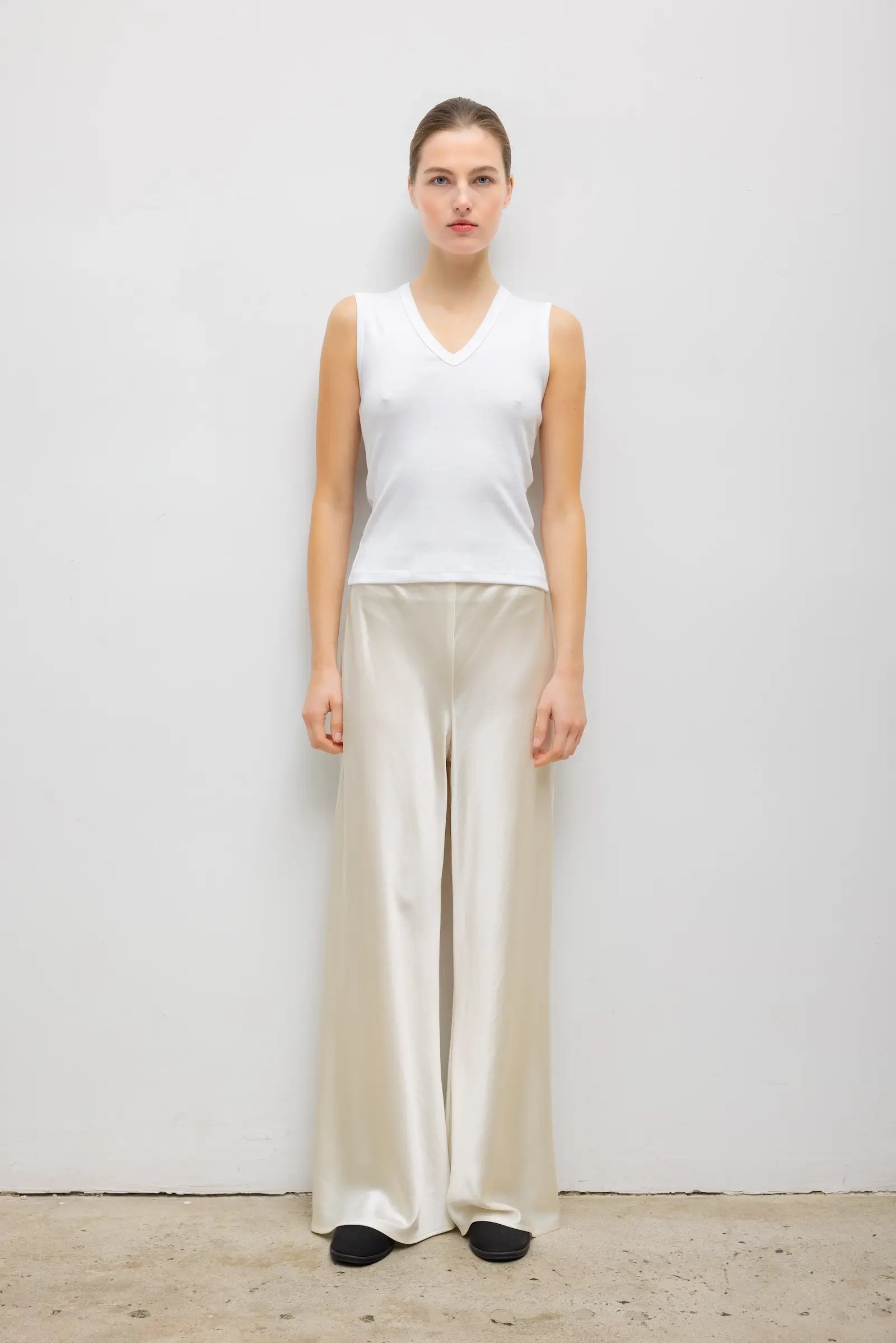 A woman stands against a plain light gray wall wearing a sleeveless white V-neck top and wide-leg satin-like pants in a light beige or cream color, with black shoes.