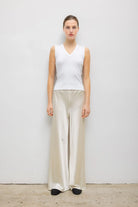 A woman stands against a plain light gray wall wearing a sleeveless white V-neck top and wide-leg satin-like pants in a light beige or cream color, with black shoes.