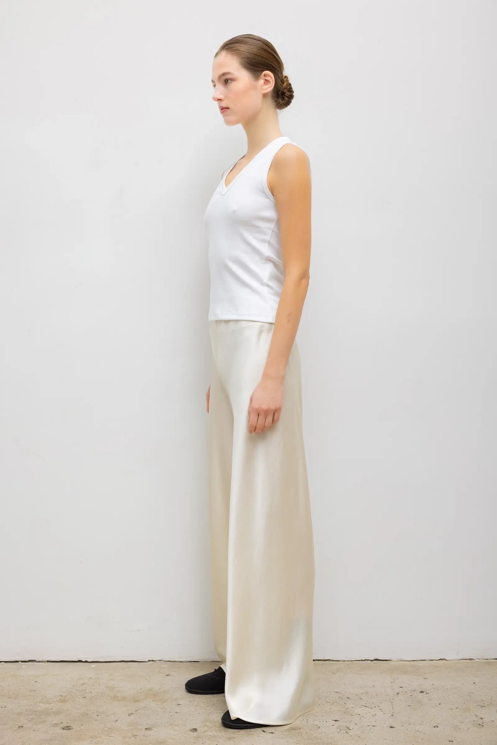 A woman stands in profile wearing a white sleeveless top and flowing cream satin wide-leg pants against a plain light gray wall.