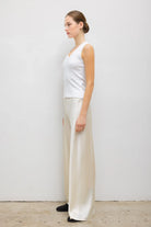A woman stands in profile wearing a white sleeveless top and flowing cream satin wide-leg pants against a plain light gray wall.