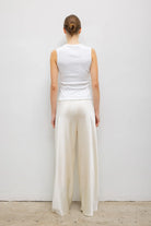 A woman viewed from behind wearing a white sleeveless top and cream wide-leg trousers, standing against a plain light gray wall.