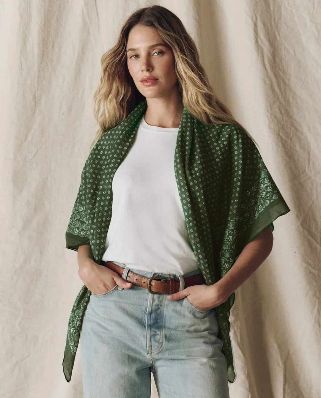 A woman models a dark green patterned shawl draped over her shoulders, worn over a white t-shirt and light-wash jeans with a brown belt, set against a neutral textured beige backdrop.