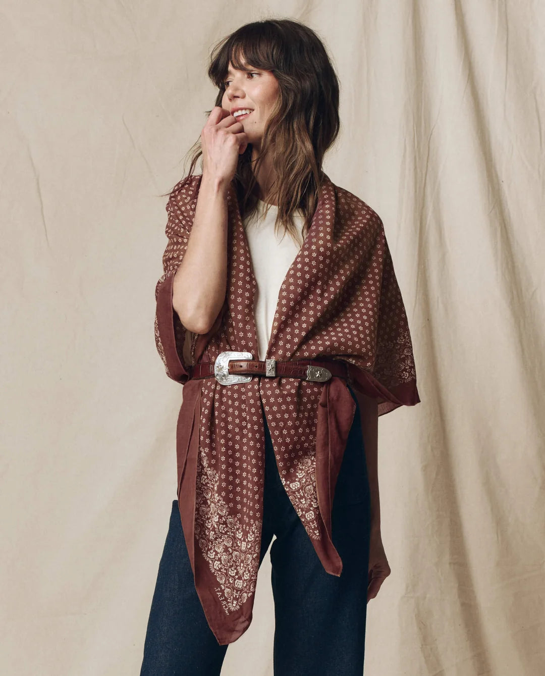 A woman models a rust-brown patterned shawl draped over her shoulders, tied at the waist with a brown belt, worn over a white top and dark blue jeans against a beige fabric backdrop.