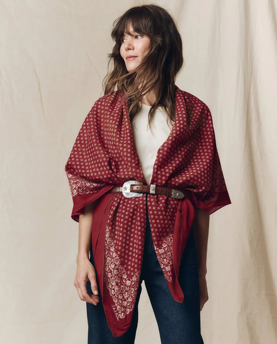 A woman wearing a red bandana-patterned wrap shawl tied around her waist with a brown belt, styled over a white top and dark blue jeans against a neutral beige fabric backdrop.