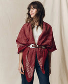 A woman wearing a red bandana-patterned wrap shawl tied around her waist with a brown belt, styled over a white top and dark blue jeans against a neutral beige fabric backdrop.