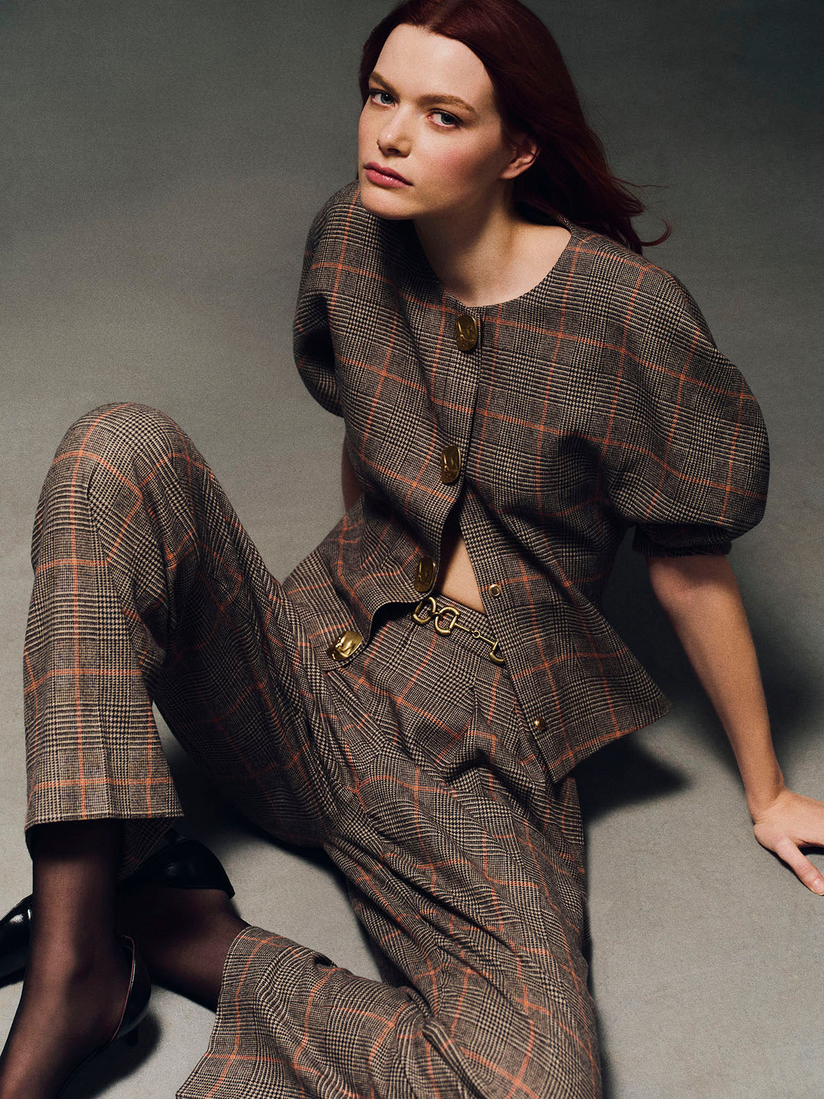 A model with red hair sits on a gray studio floor wearing a brown and beige plaid matching set consisting of a short-sleeve cropped jacket with gold-tone buttons and wide-leg trousers.