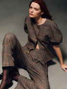 A model with red hair sits on a gray studio floor wearing a brown and beige plaid matching set consisting of a short-sleeve cropped jacket with gold-tone buttons and wide-leg trousers.