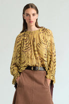 A woman wearing a yellow and brown patterned long-sleeve blouse tucked into a brown skirt with a black belt, photographed against a light gray studio background.