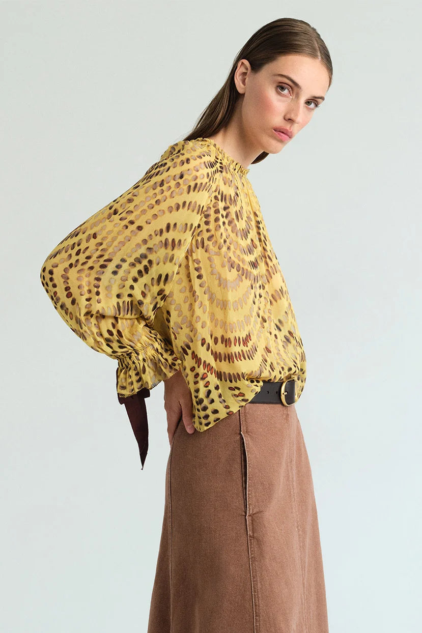 A woman modeling a yellow and brown leopard-print sheer blouse with long balloon sleeves, paired with a brown skirt, against a light gray studio background.