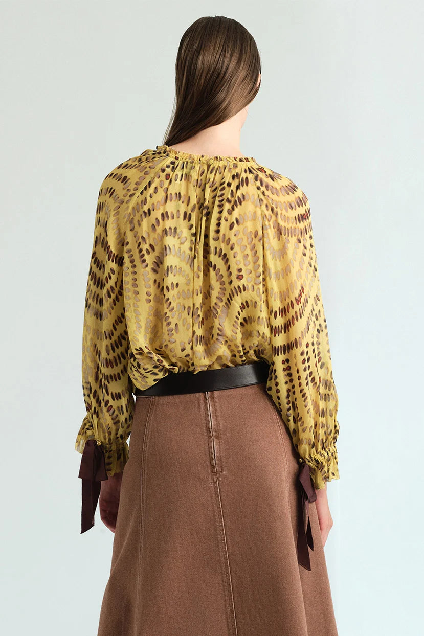 A model shown from the back wearing a yellow and brown patterned long-sleeve blouse tucked into a brown skirt with a black belt, against a light gray studio background.