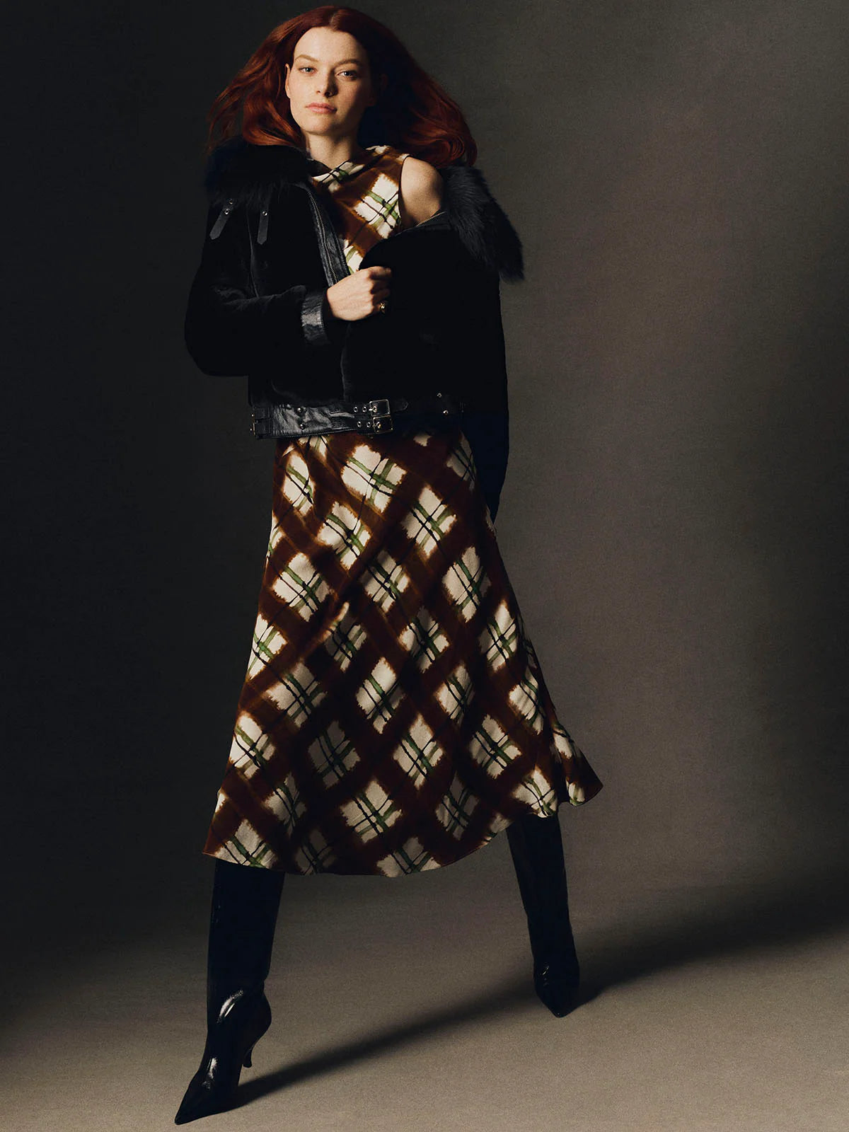 A model with red hair poses in a studio wearing a black leather jacket with dark fur trim, a brown and beige plaid midi skirt, and black pointed-toe heels against a dark gray background.