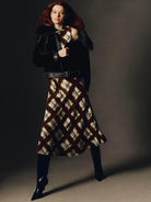 A model with red hair poses in a studio wearing a black leather jacket with dark fur trim, a brown and beige plaid midi skirt, and black pointed-toe heels against a dark gray background.