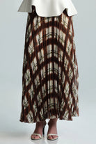 A pleated maxi skirt with a brown and beige shibori tie-dye pattern, styled with a white ruffled top and beige heeled sandals against a light gray background.