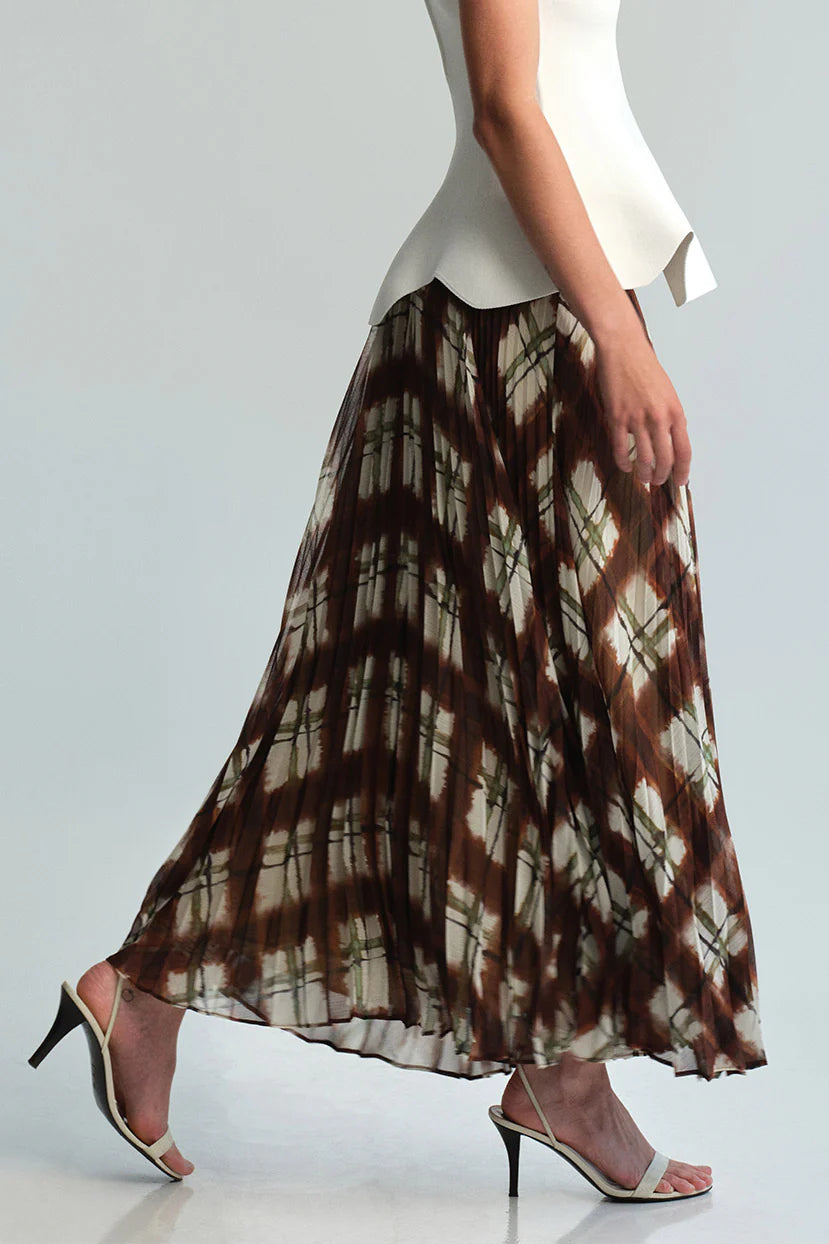 A woman wearing a long pleated maxi skirt with a brown and beige tie-dye diamond pattern, paired with a white peplum top and white heeled sandals, shown from the waist down against a light gray studio background.
