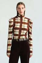 A model wearing a brown and cream plaid mock neck long-sleeve top with puffed shoulders, paired with dark brown trousers and a black belt, against a light gray studio background.