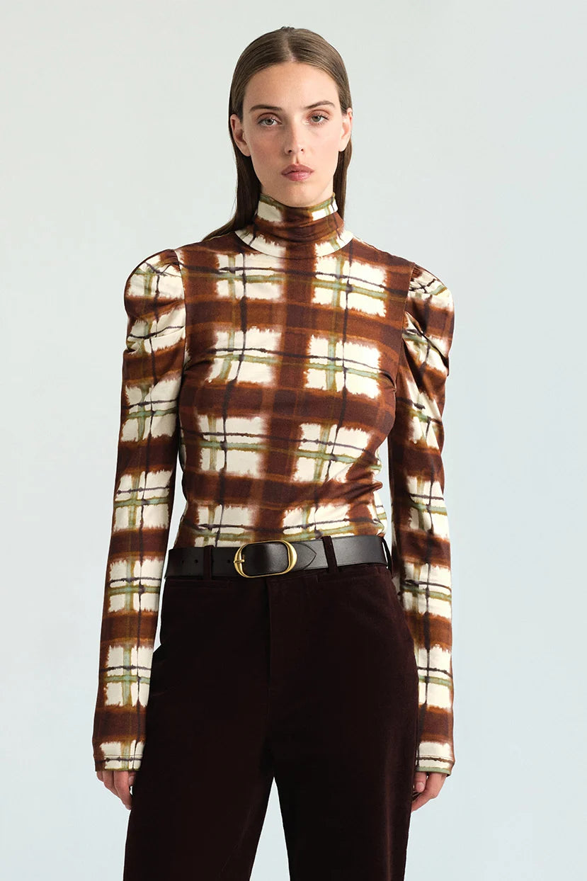 A model wearing a brown and cream plaid mock neck long-sleeve top with puffed shoulders, paired with dark brown trousers and a black belt, against a light gray studio background.