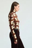 A woman shown in side profile wearing a fitted mock-neck long-sleeve top with a brown, beige, and white plaid pattern, paired with dark brown pants and a black belt, against a light gray studio background.
