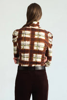 A model wearing a brown and cream plaid mock neck long-sleeve top with puffed shoulders, paired with dark brown trousers and a black belt, against a light gray studio background.
