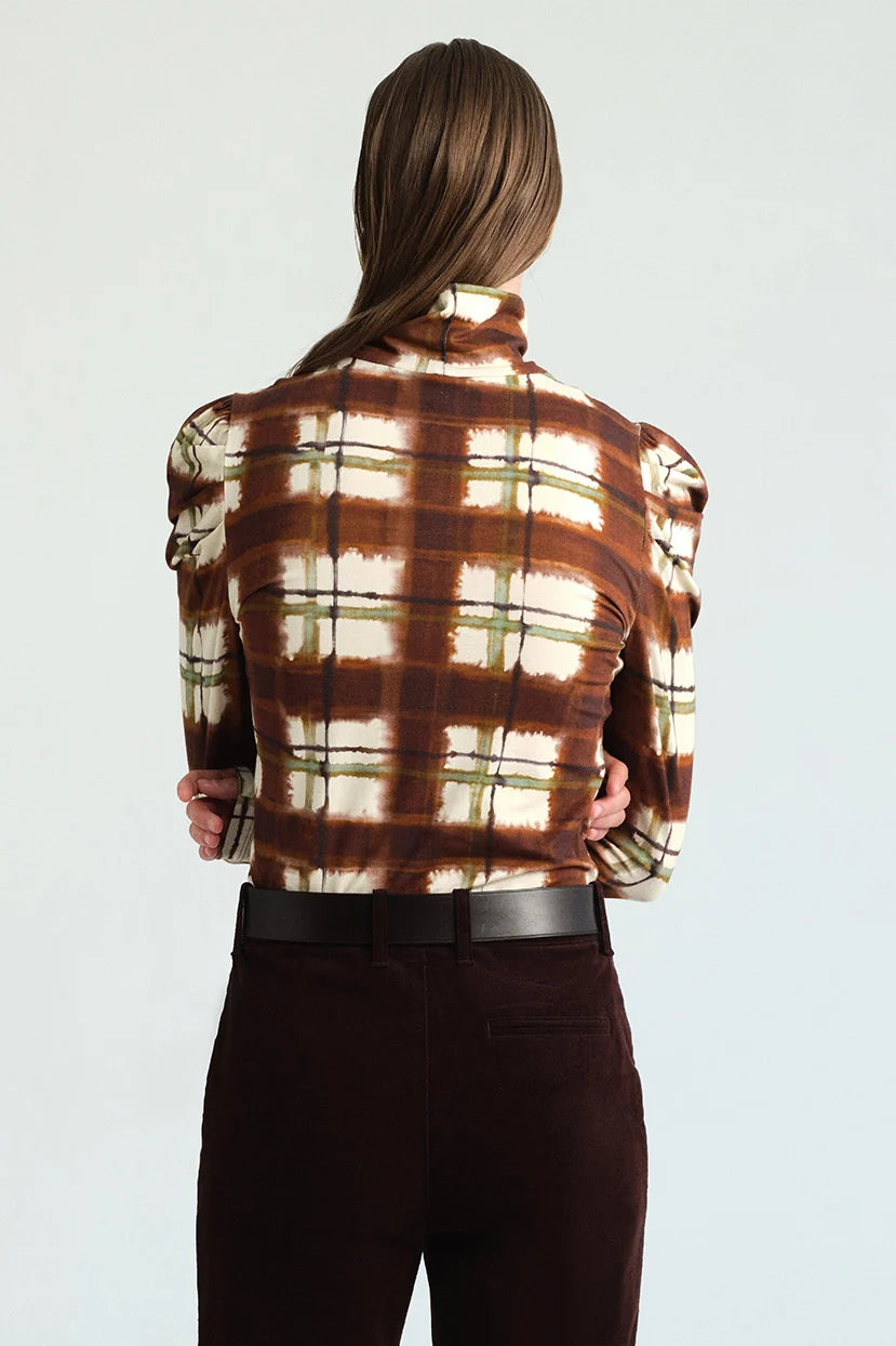 A model wearing a brown and cream plaid mock neck long-sleeve top with puffed shoulders, paired with dark brown trousers and a black belt, against a light gray studio background.
