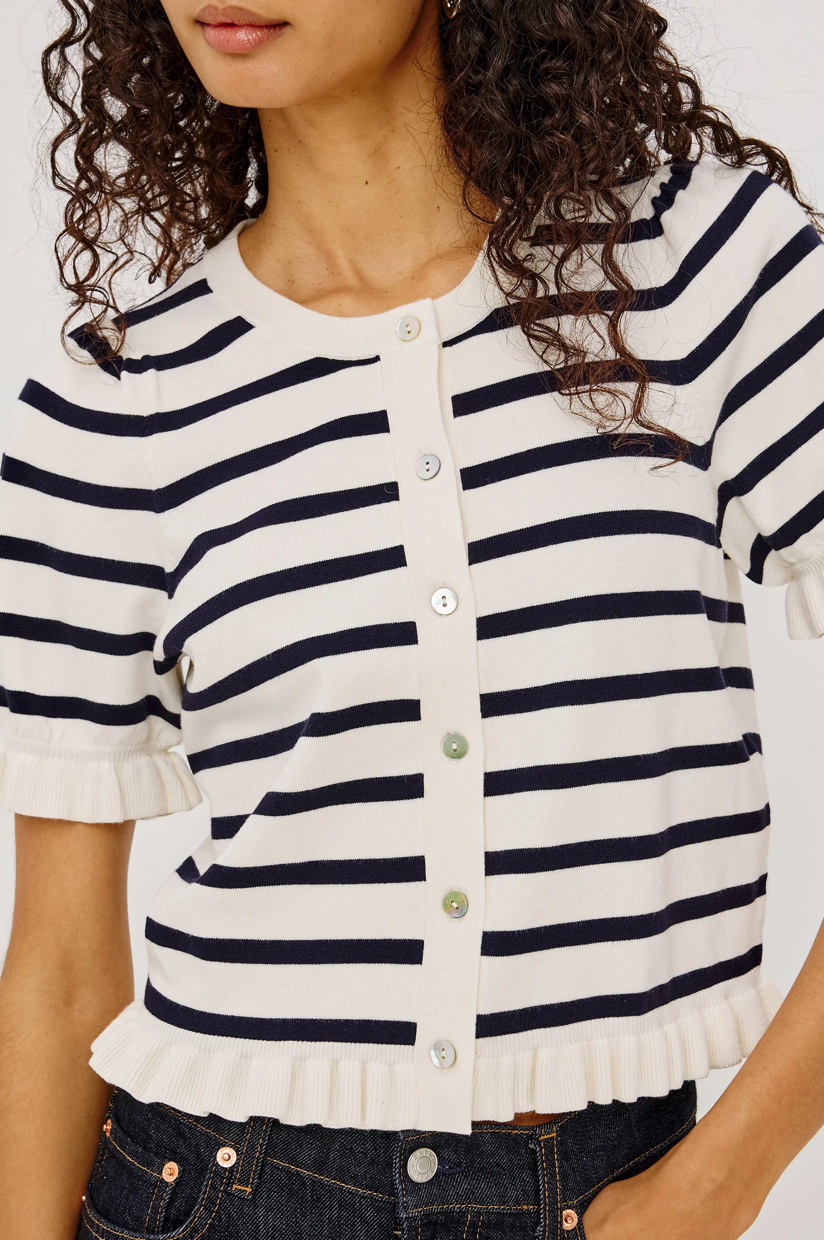 A close-up of a woman wearing a short-sleeve knit top with horizontal navy and off-white stripes, a round neckline, and a button-front closure with ruffled trim at the sleeves and hem.