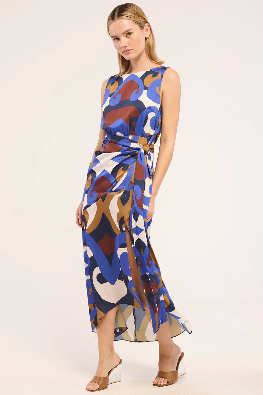 A woman modeling a sleeveless midi dress with an abstract multicolor print in blue, brown, beige, and white, featuring a tie waist and side slit, set against a plain light background.