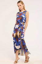 A woman modeling a sleeveless midi dress with an abstract multicolor print in blue, brown, beige, and white, featuring a tie waist and side slit, set against a plain light background.