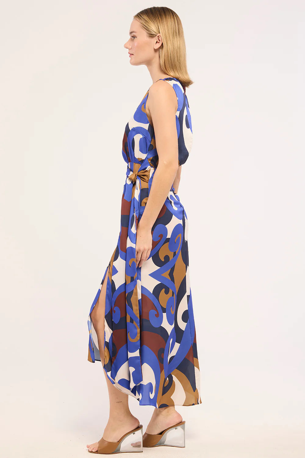 A woman modeling a sleeveless midi dress with a blue, brown, and white abstract pattern, shown from a side profile against a plain light background.