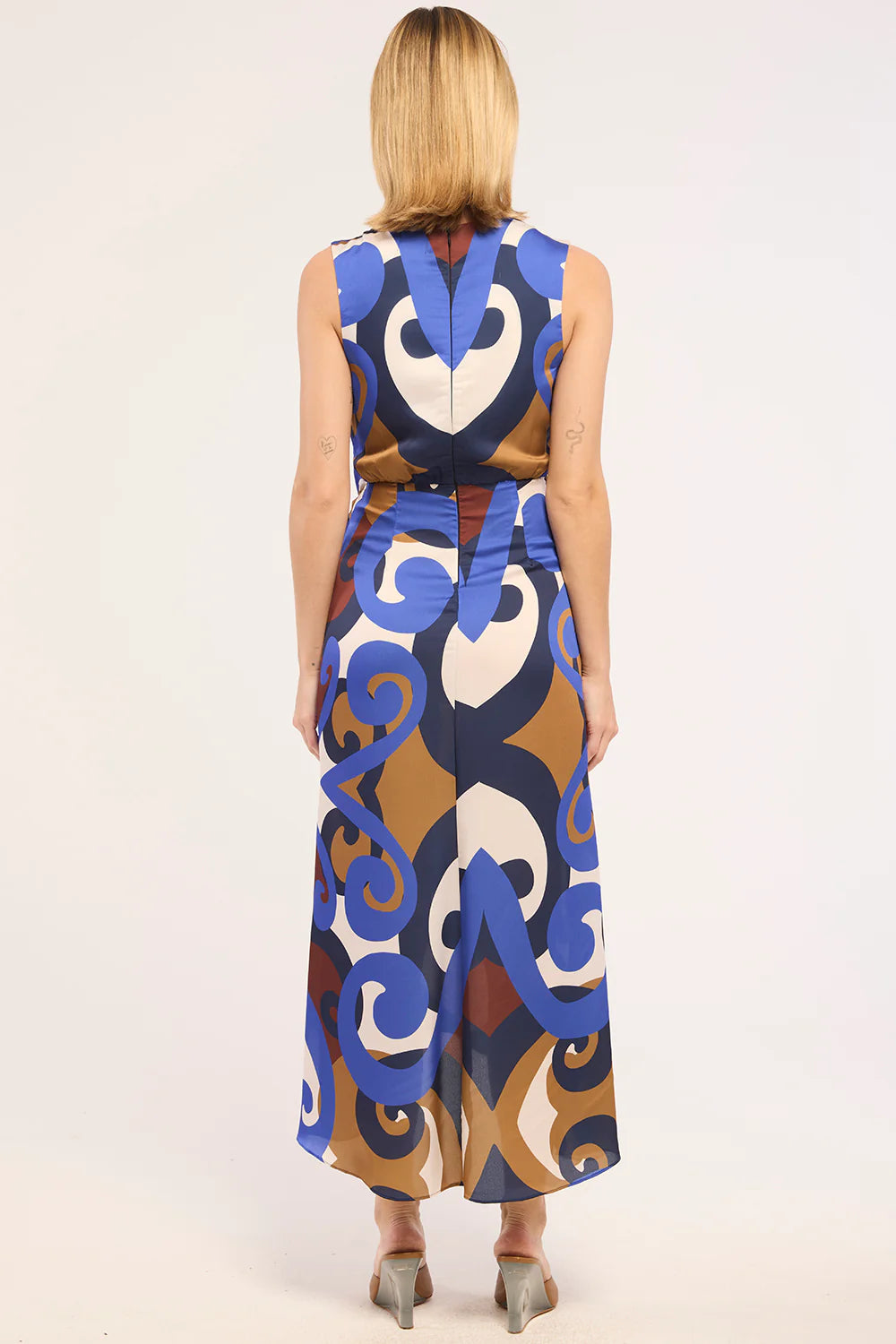 A woman viewed from the back wearing a sleeveless midi dress with a bold abstract pattern in blue, brown, cream, and white, paired with gray heels, against a plain light background.