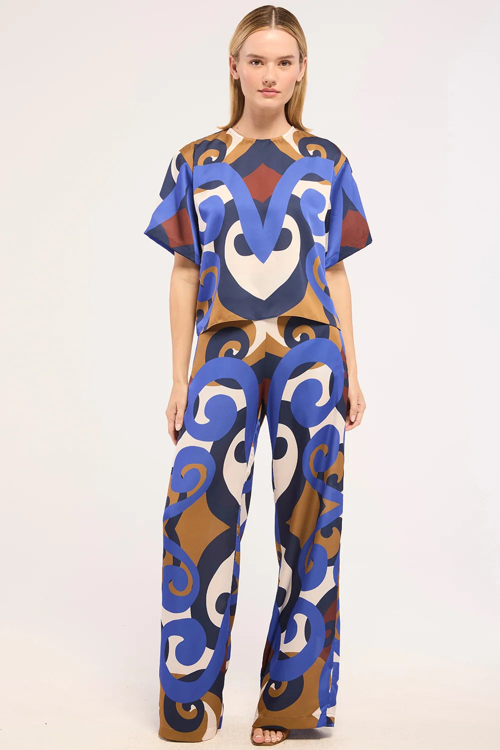 A woman modeling a short-sleeve jumpsuit with a bold abstract pattern in blue, brown, beige, and white against a plain light background.