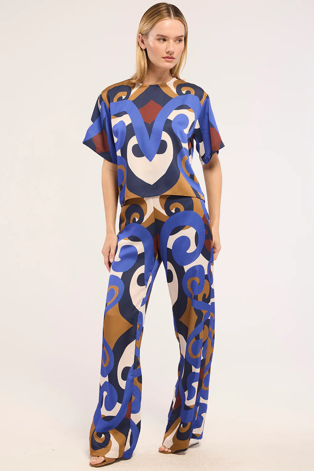 A woman modeling a matching short-sleeve top and wide-leg pants set with a bold abstract pattern in blue, brown, cream, and burgundy on a light gray studio background.