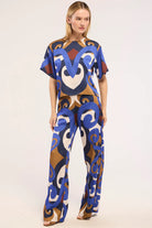 A woman modeling a matching short-sleeve top and wide-leg pants set with a bold abstract pattern in blue, brown, cream, and burgundy on a light gray studio background.