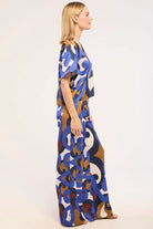 A woman modeling a short-sleeved maxi dress with an abstract, painterly pattern in blue, brown, beige, and white, shown from a side profile against a plain white studio background.