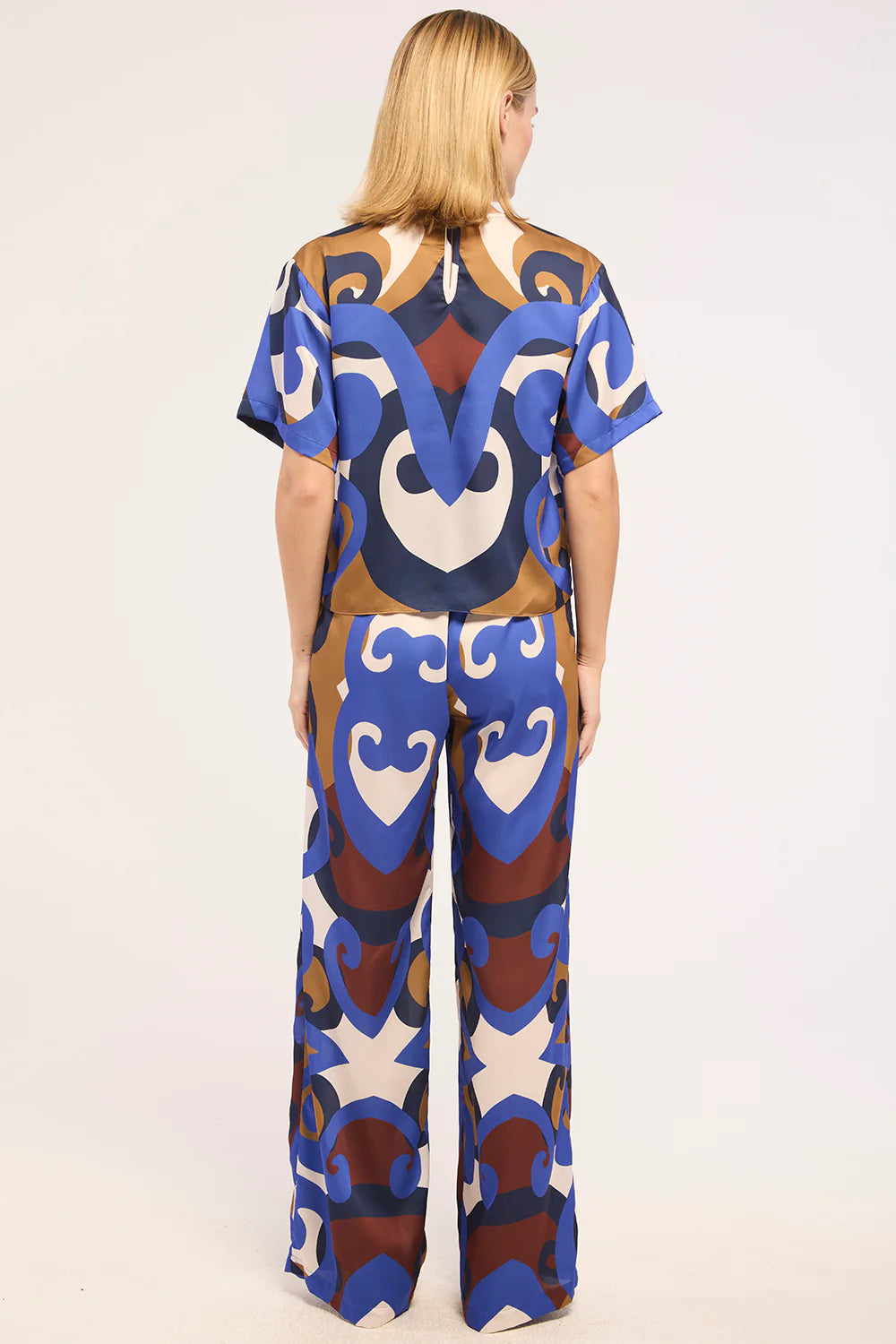 A woman viewed from behind wearing a matching short-sleeve top and wide-leg pants set with a bold abstract pattern in blue, brown, beige, and white on a light gray studio background.
