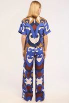 A woman viewed from behind wearing a matching short-sleeve top and wide-leg pants set with a bold abstract pattern in blue, brown, beige, and white on a light gray studio background.