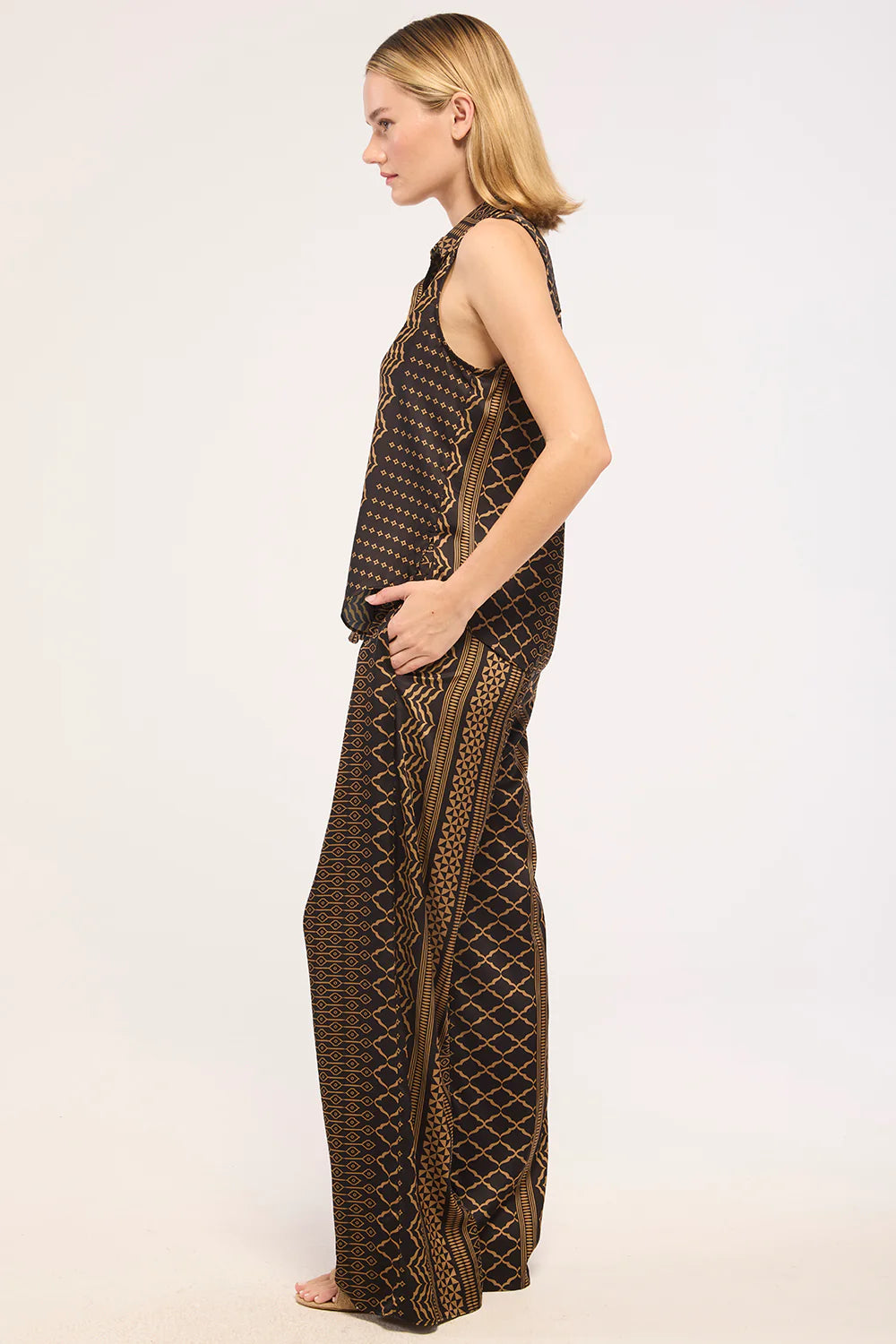 A woman in profile wearing a sleeveless brown and black patterned maxi dress with geometric designs, standing against a plain white studio background.