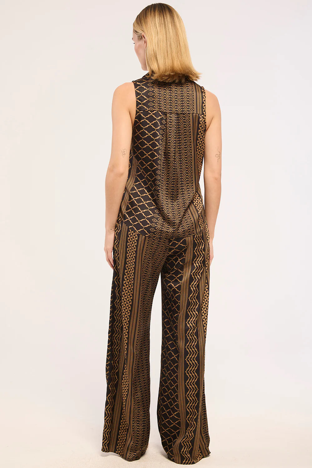 A woman viewed from the back wearing a sleeveless brown and black patterned jumpsuit with wide legs, photographed against a plain light background.