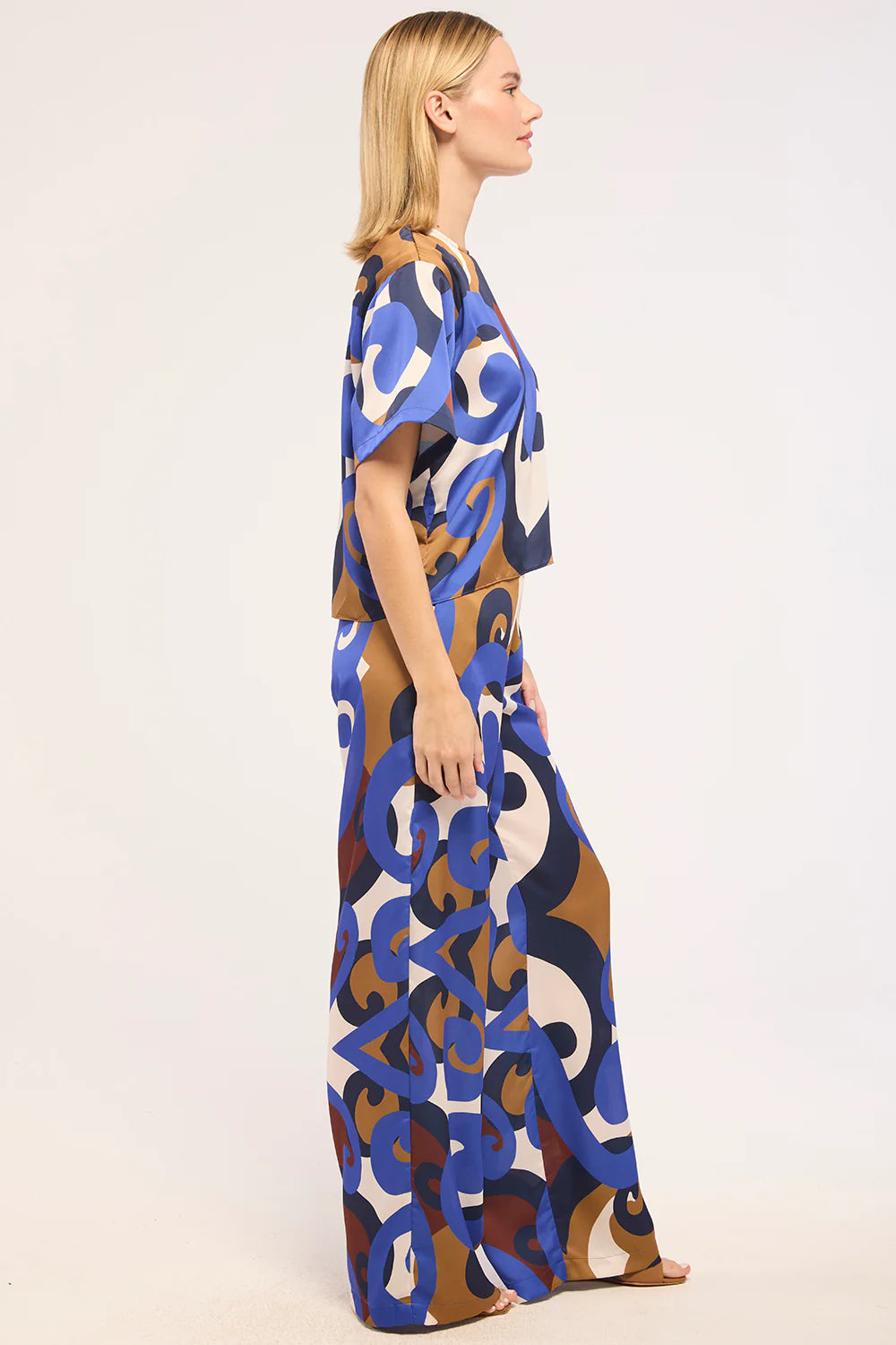 A woman modeling a short-sleeved maxi dress with an abstract, painterly pattern in blue, brown, beige, and white, shown from a side profile against a plain white studio background.